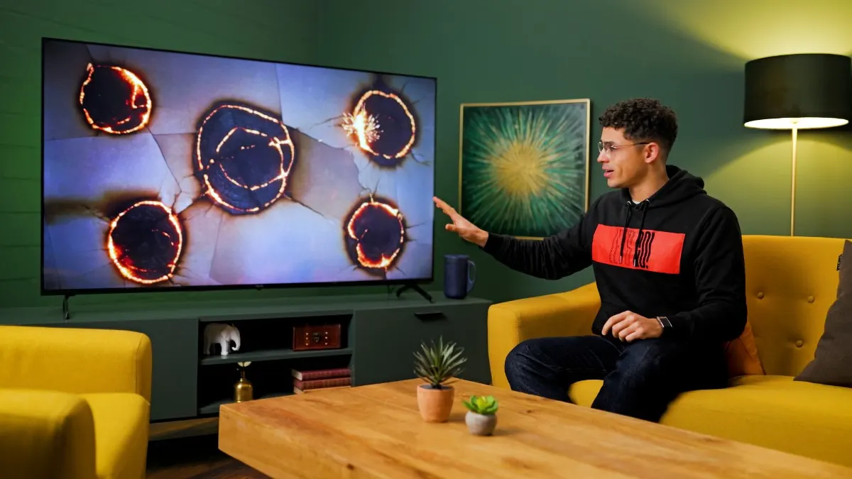 TURN ON Moderator Ilias sitzt auf einer gelben Couch und zeigt mit ausgestreckter Hand auf einen TV mit simulierten Einbrenneffekten.