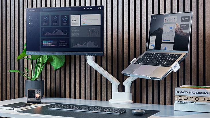 Ein Schreibtisch mit Monitor, Laptop auf Halterung, Tastatur, Pflanze und Lautsprecher vor einer Holzpaneelwand.