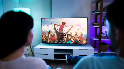 Zwei Personen schauen auf einen Fernseher, der eine Menge jubelnder Menschen zeigt, Konfetti fällt. Ein Regal steht rechts.