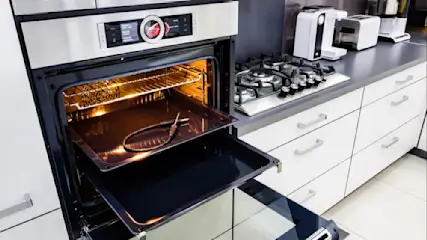 Offener, beleuchteter Backofen mit Blech und Zubehör. Im Hintergrund Herd, Toaster und weiße Küchenschränke.