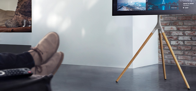 Ein Fernseher steht auf einem Stativ vor einer Ziegelwand. Füße in Schuhen liegen auf einem Tisch mit einer Fernbedienung.