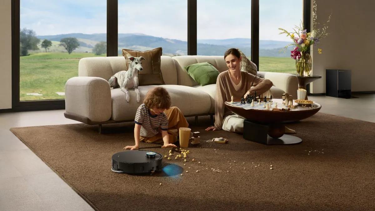 Eine Frau und ein kleiner Junge sitzen mit einem Hund in einem Wohnzimmer, während der Dreame X60 Pro Ultra Complete den Teppich saugt.
