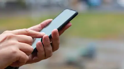 Eine Person hält ein schwarzes Smartphone in der Hand und tippt mit dem Finger auf den Bildschirm, im Hintergrund ist eine unscharfe grüne Fläche.