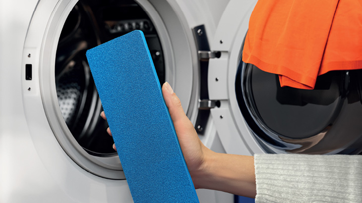 Offene Waschmaschine mit orangefarbener Wäsche. Eine Hand hält einen blauen Pad vor der geöffneten Waschmaschine. 