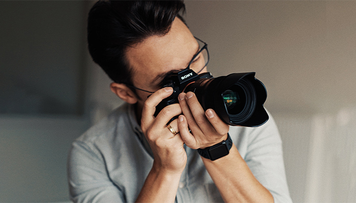 Ein Mann mit Brille hält eine Sony-Kamera mit einem schwarzen Objektiv vor sein Gesicht. 