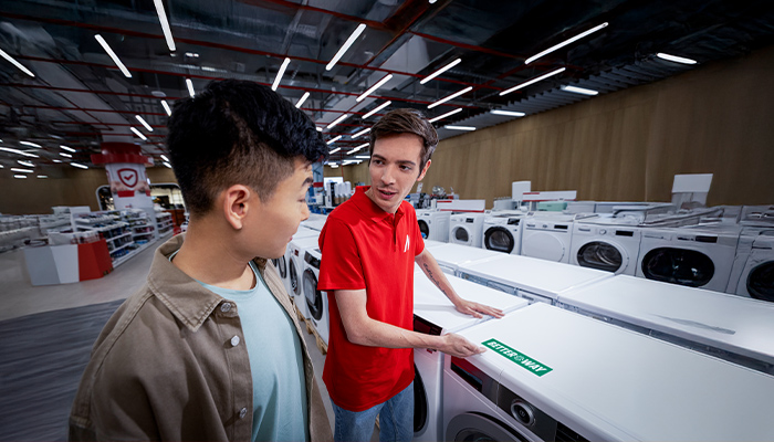 In einem Geschäft stehen zwei Männer vor einer Reihe von Waschmaschinen. Einer trägt ein rotes Hemd, der andere eine Jacke.