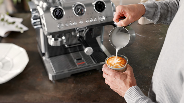 Person gießt feinporigen Milchschaum in einen Latte Art Kaffee neben einer modernen Siebträgermaschine.