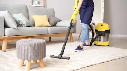Eine Person mit gelben Handschuhen staubsaugt einen Teppich. Im Hintergrund befinden sich ein graues Sofa, ein Hocker und ein gelber Staubsauger.