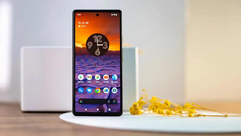 Auf einem Tisch steht ein Smartphone mit einem benutzerdefinierten Startbildschirm vor einem weißen Objekt und gelben Blumen.