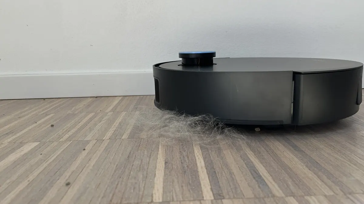 Ein schwarzer Staubsaugerroboter steht auf einem Holzboden vor einer weißen Wand und saugt Staub und Fusseln auf.