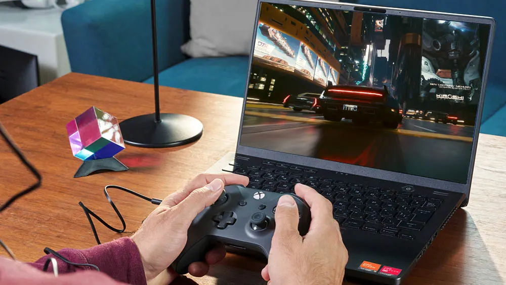 Ein Laptop zeigt ein Videospiel, das mit einem Xbox-Controller auf einem Holztisch gespielt wird. Ein Würfel und eine Lampe stehen daneben.