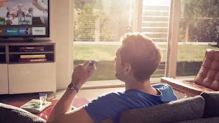 Ein Mann sitzt auf einer Couch mit Fernbedienung. Ein Fernseher, ein Fenster und ein Sessel sind im Raum.