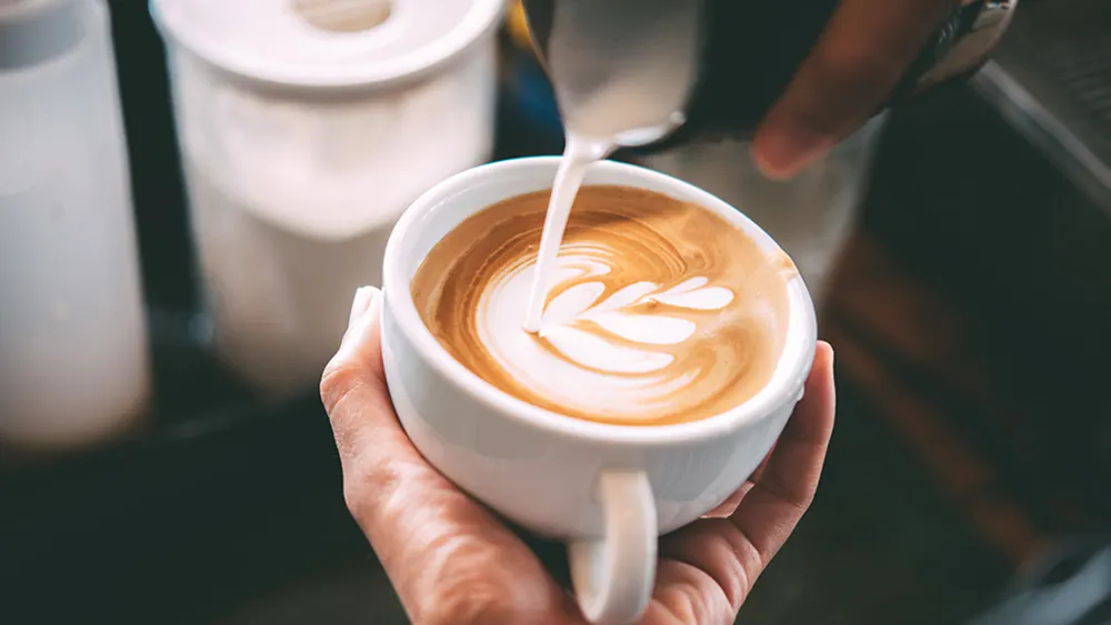 Eine Hand hält eine weiße Tasse mit Latte Art, während Milch hineingegossen wird. Weiße Behälter stehen im Hintergrund.
