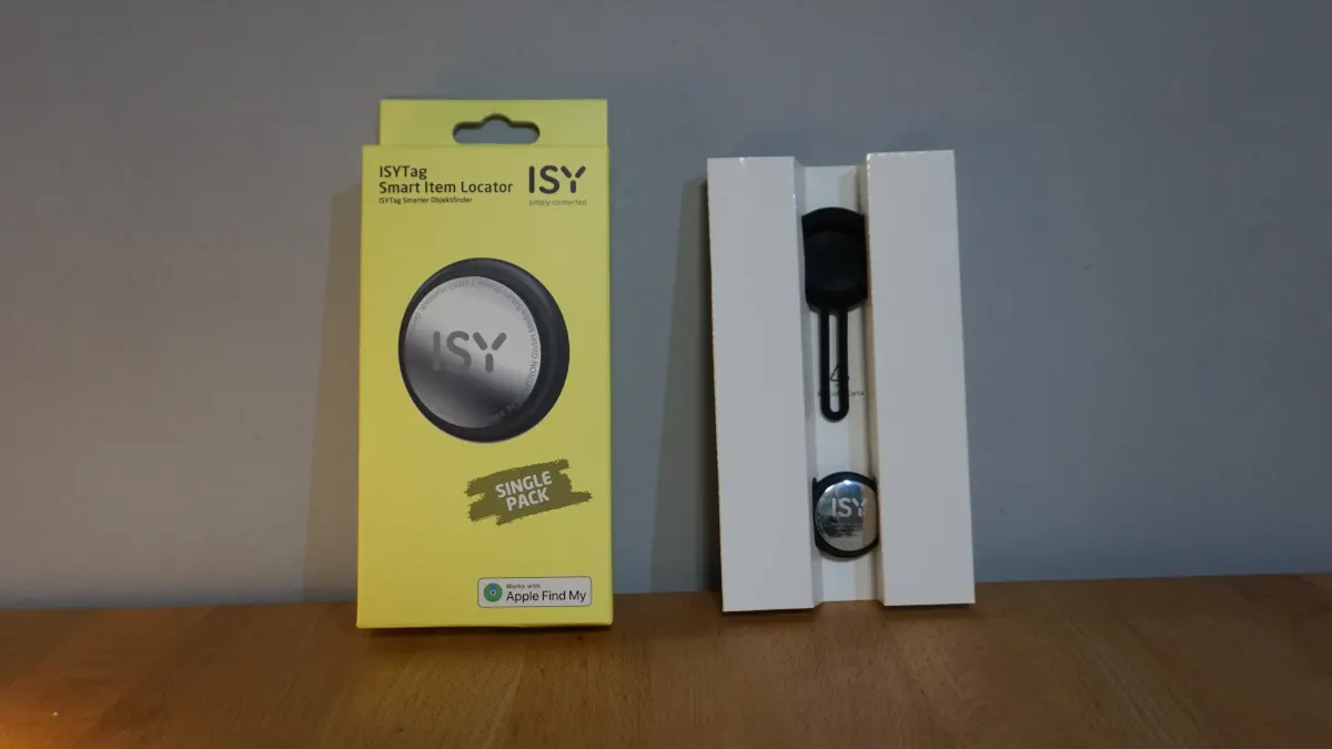 Gelbe ISYTag Einzelpackung und weiße Verpackung mit schwarzem Schlüsselanhänger auf einem Holztisch vor einer grauen Wand.