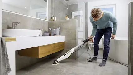 Ein Mann reinigt ein Badezimmer mit einem weißen Dampfreiniger auf grauen Fliesen. Waschbecken, Spiegel und Badewanne sind im Hintergrund.