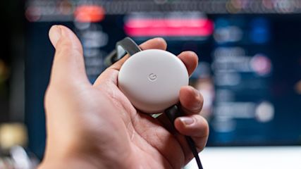 Ein Mann hält einen Google Chromecast in der Hand, im Hintergrund ist ein TV zu sehen.
