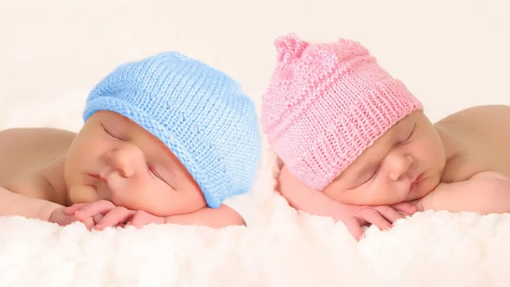 Zwei Babys, eines mit blauer, das andere mit rosa Strickmütze, schlafen friedlich auf einer weichen, weißen Decke.