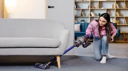 Eine Frau saugt mit einem Akku-Staubsauger den Teppich unter einem Sofa.