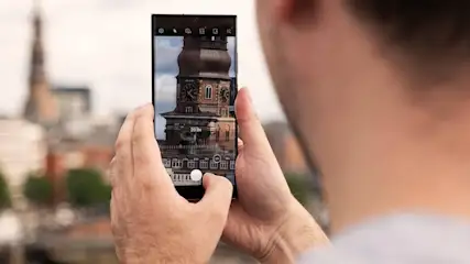 Mann hält ein Smartphone, das einen Turm mit einer Uhr anzeigt. Ein weiterer Turm ist im Hintergrund zu sehen.