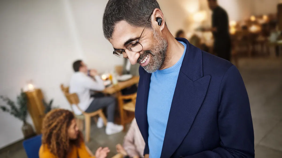 Ein Mann mit Brille und Kopfhörer, trägt einen blauen Pullover und Blazer. Im Hintergrund sitzen Personen an Tischen.
