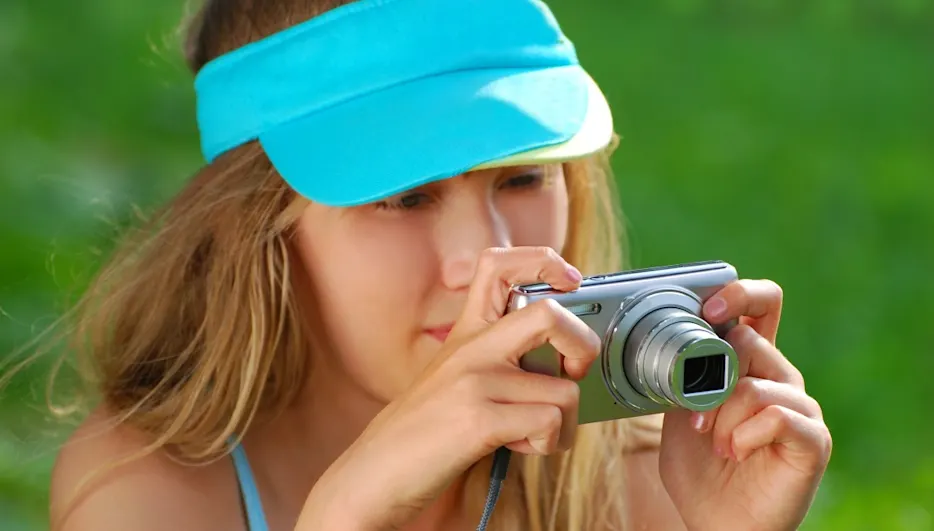 Frau mit blauem Visier hält eine silberne Kamera vor einem grünen Hintergrund.