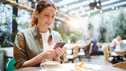 Frau mit olivgrünem Hemd benutzt Smartphone an Tisch mit Kaffee und Gebäck, Pflanzen und andere Personen im Hintergrund.