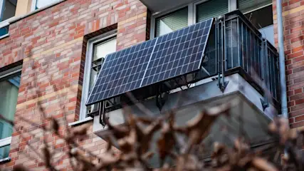 Ein Solarpanel ist an einem Balkon mit Metallgeländer eines Backsteingebäudes befestigt. Fenster sind sichtbar.