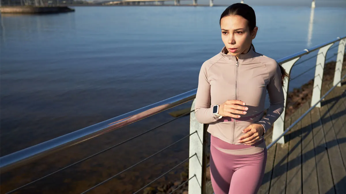 Frau in Sportkleidung mit Kopfhörern blickt auf eine blaue Smartwatch vor einer Stadtkulisse und einem Geländer.