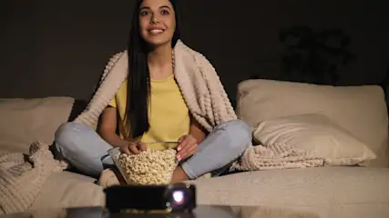 Eine Frau sitzt mit einer Decke auf einem Sofa, hält Popcorn und schaut einen Film, der von einem Projektor gezeigt wird.