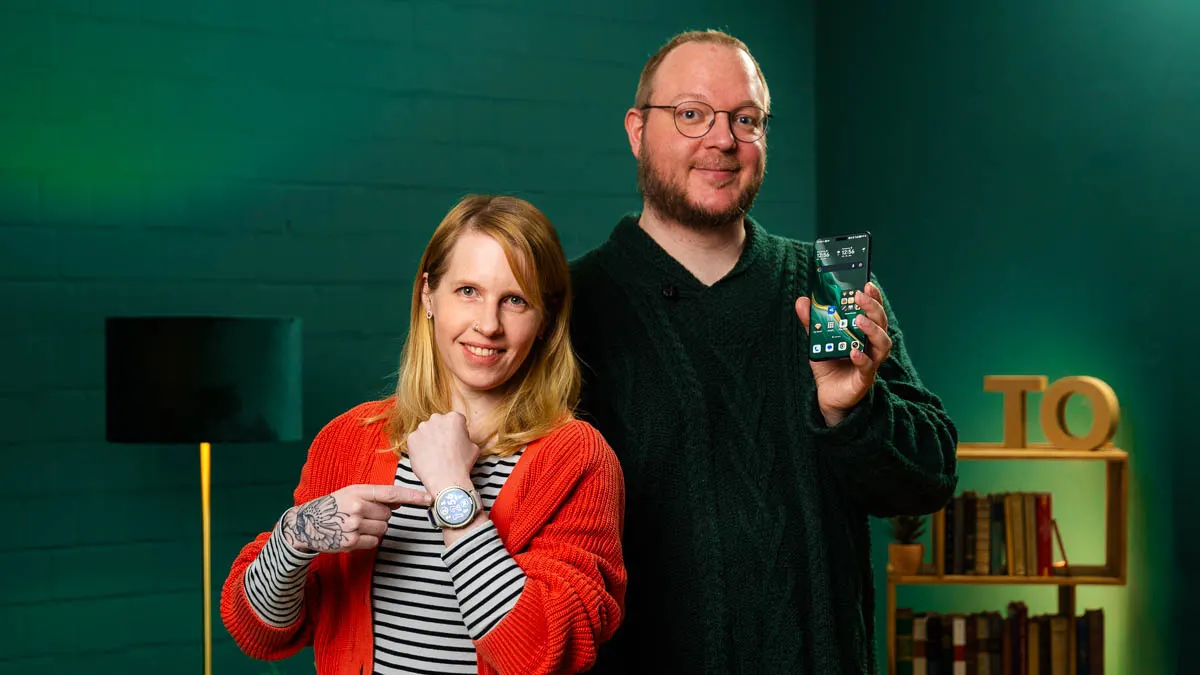 Franzi zeigt auf ihre Smartwatch, Andreas hält sein Smartphone hoch.