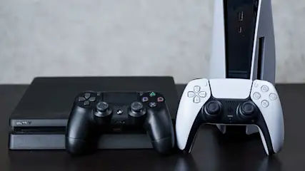 Auf einem Tisch stehen eine schwarze PS4, eine weiße PS5 und zwei Controller in Schwarz und Weiß.