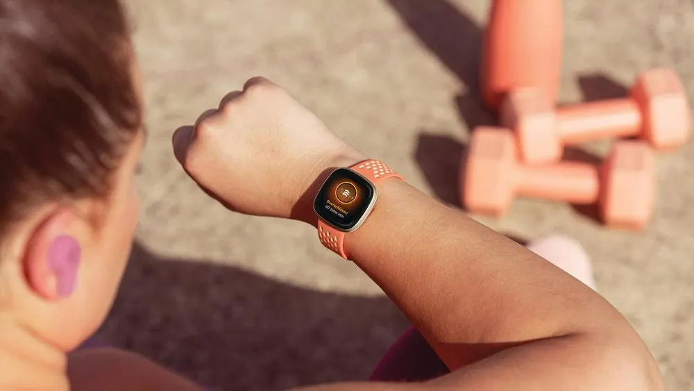Nahaufnahme einer Person mit Smartwatch am Handgelenk und Hanteln im Hintergrund. Die Uhr hat ein oranges Armband.