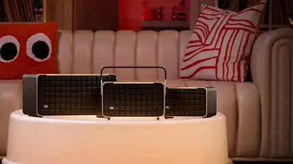 Drei schwarze JBL-Lautsprecher mit Goldrand auf einem runden, weißen Tisch, im Hintergrund ein beiges Sofa mit Kissen.
