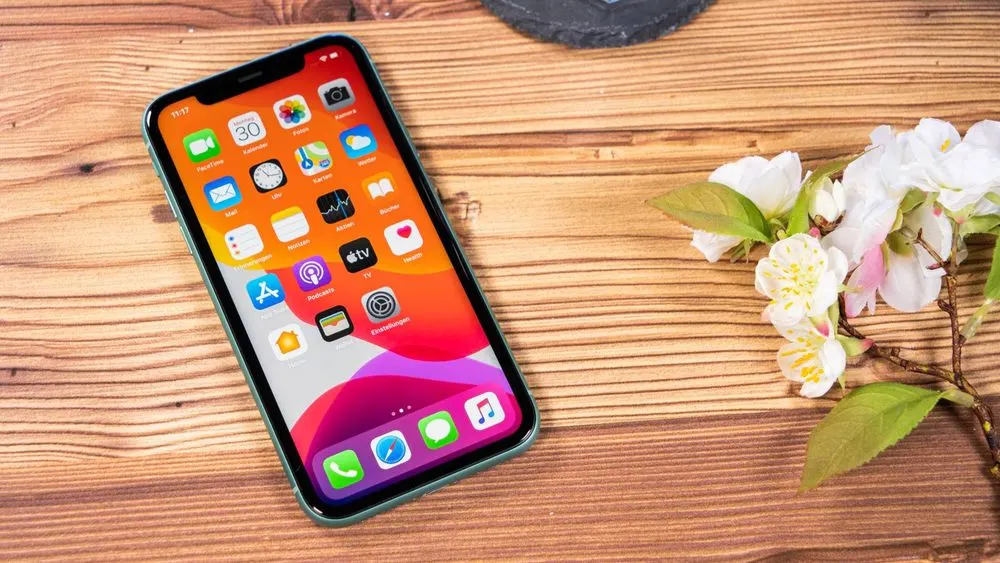 Ein Smartphone mit bunten App-Symbolen liegt auf einem Holztisch, daneben weiße Blüten mit grünen Blättern.