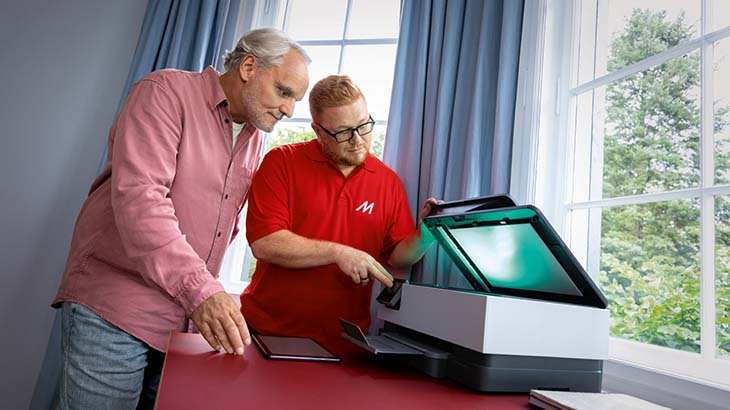 Zwei Männer stehen an einem roten Tisch neben einem Drucker vor einem Fenster mit Bäumen.