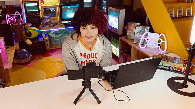 Eine Person sitzt vor einem Laptop und einer Kamera auf einem Tisch, im Hintergrund Regale und Bildschirme.