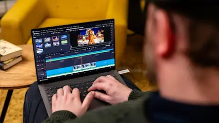 Eine Person bearbeitet ein Video auf einem Laptop, der auf ihren Knien liegt. Ein gelber Sessel und ein Tisch mit Büchern sind sichtbar.