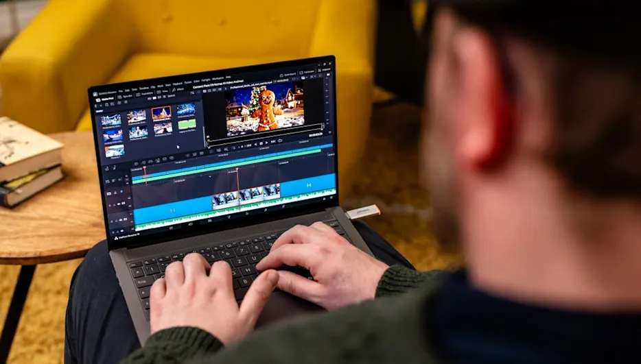 Eine Person bearbeitet ein Video auf einem Laptop, der auf ihren Knien liegt. Ein gelber Sessel und ein Tisch mit Büchern sind sichtbar.
