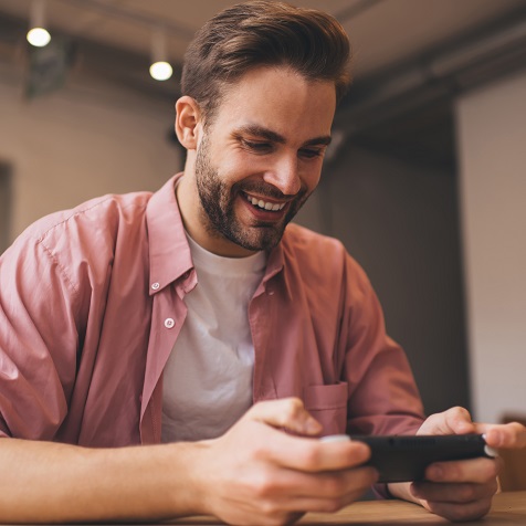 Ein Mann lächelt und hält ein Smartphone horizontal vor sich. Er trägt ein rosa Hemd und ein weißes T-Shirt.