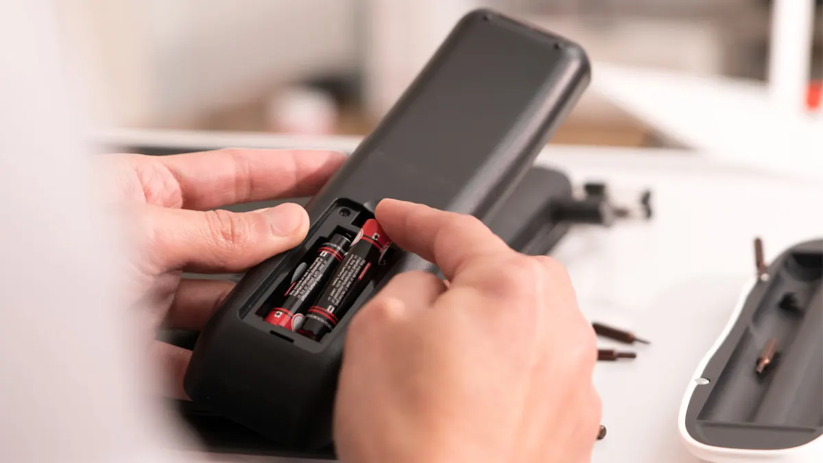 Eine Hand legt eine Batterie über ein Batteriefach einer Fernbedienung.