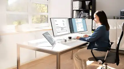 Eine Frau sitzt an einem Schreibtisch mit zwei Monitoren und einem Laptop in einem hellen Büro.