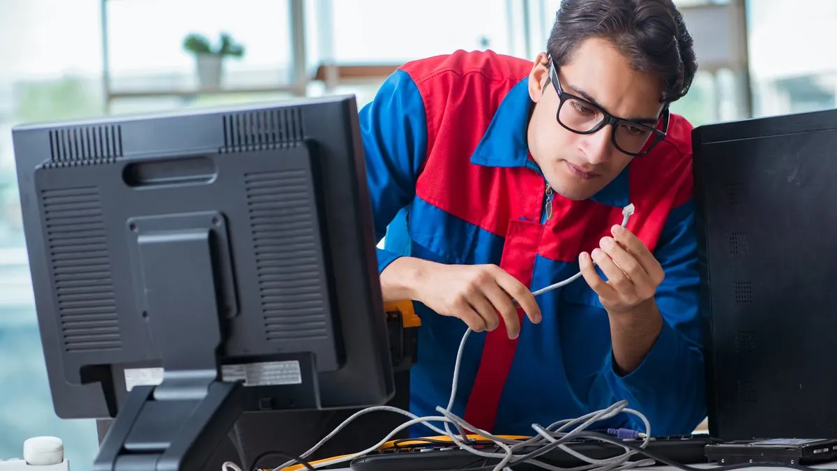 Ein Mann mit Brille, in blau-roter Arbeitskleidung, hält ein weißes Kabel vor schwarzen Computermonitoren und Kabeln.