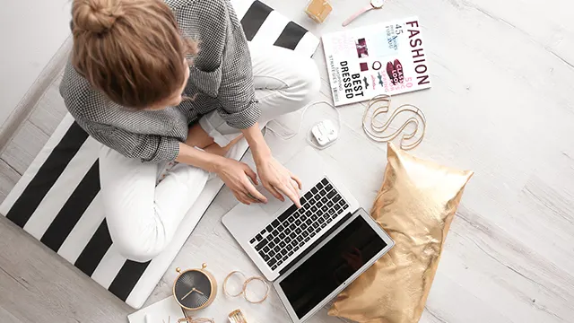 Eine Frau sitzt mit einem Laptop auf dem Boden, umgeben von Kissen, einer Zeitschrift, einer Uhr und anderen Gegenständen.