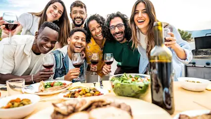 Eine Gruppe von acht Personen lacht und hält Weingläser hinter einem Tisch mit Speisen, darunter Salat und Fleisch.