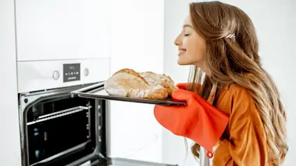 Eine Frau hält ein Backblech mit frisch gebackenem Brot, im Hintergrund ein geöffneter Backofen.