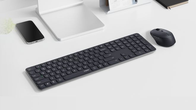Schwarze Tastatur und passende Maus auf einem aufgeräumten weißen Schreibtisch; daneben liegen ein Smartphone, ein Monitorständer und ein Notizbuch im minimalistischen Arbeitsumfeld.