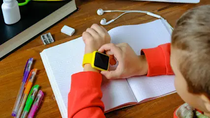 Ein Kind mit rotem Pullover bedient eine gelbe Smartwatch über einem offenen Notizbuch. Stifte, Kopfhörer und Bürobedarf liegen auf einem Tisch.