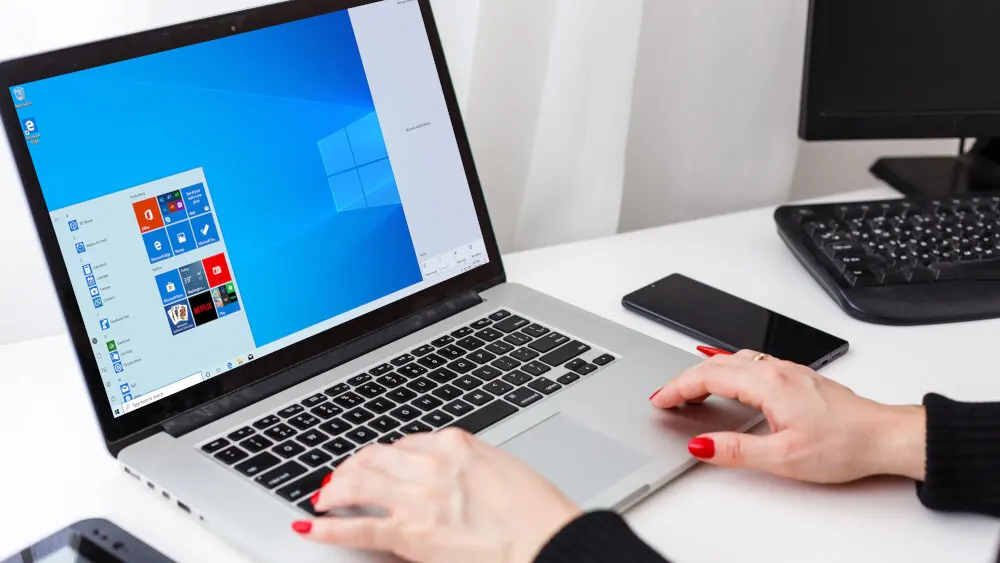 Eine Person tippt auf einem Laptop mit blauem Bildschirm. Ein schwarzes Handy und eine schwarze Tastatur liegen auf einem weißen Tisch.
