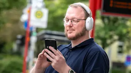 Ein Mann mit Brille und Kopfhörern hält ein Smartphone in der Hand. Im Hintergrund sind Bäume und ein Buswartehäuschen zu sehen.