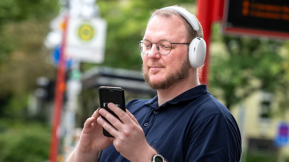 Ein Mann steht an einer Bushaltestelle während er ein Smartphone in den Händen hält und über Kopfhörer Musik hört.